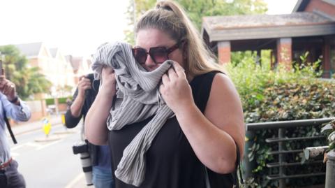 A woman holds clothing to her face to obscure it from view, she is wearing large dark sunglasses, has her hair tied back and is wearing a sleeveless back top. She looks distressed.