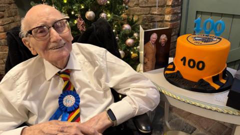 A 100-year-old man smiles as he sits next to a table with a birthday cake and a card showing a picture of the King and Queen. The cake is amber and black, with a Hull City logo on the top and candles spelling out "100". The man is wearing a smart white shirt, a tie in the colours of Hull City and a rosette badge that reads "birthday boy". A Christmas tree stands in the background.