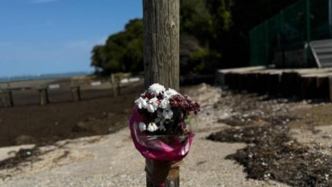 A bunch of flowers tied to a wooden stump on the beach at Binstead. The sea is visible on the left and the sky is blue.