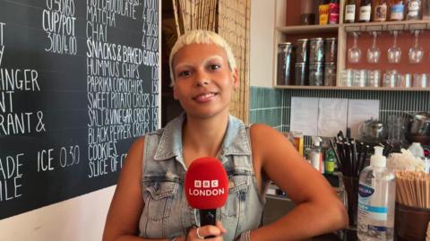 A young woman holds a red microphone as she is being interviewed. She is sitting at the counter in a cafe. 