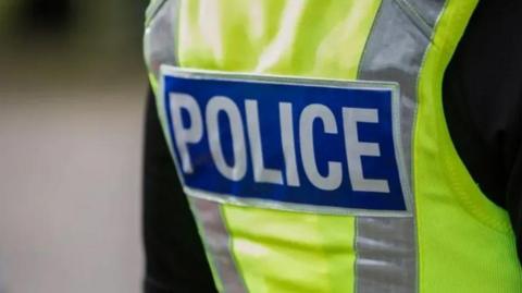 Stock, side angle shot of a yellow, hi-vis police jacket. It has 'police' written on the back in silver writing in a dark blue rectangle. There is also two vertical silver stripes, from the top of the jacket to the bottom, either side of the blue rectangle 