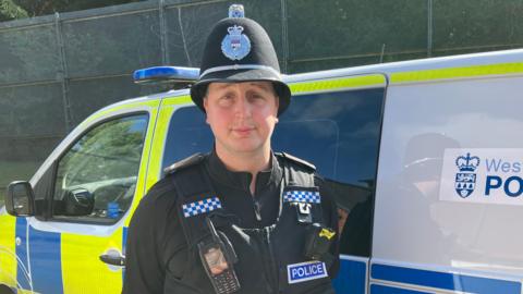 A police officer in uniform standing beside a police van.  The officer is looking seriously into the camera, he's also wearing a hat with his police radio stuck onto his jacket 