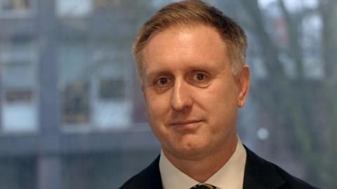 Dr Ed Garratt is smiling at the camera wearing a suit. He has short brown hair, going grey at the sides. The background is blurred it but looks like he's standing in front of a window. 