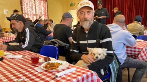 Peter wearing a black jacket and a white cap and is holding a white dog- sitting at a table with a plate of food in front 