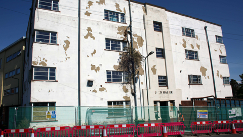 A dilapidated four-storey building. White paint is flaking off it, with windows lining he side. There are red and white striped barriers in front of it and metal railings. 