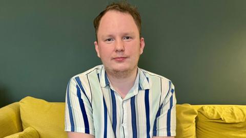 A man with a neutral expression looks at the camera. He is in his living room. He is white with brown hair.