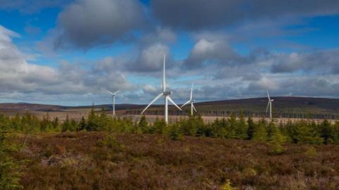 Wind turbines among trees, with hills and a cloudy blue sky