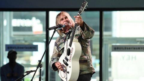 Mike Peters who has long blonde hair and is wearing a khaki camouflage jacket. He is stood at a microphone stand and is holding a cream guitar which he is playing and looking at. Behind him are some large windows and a man wearing a baseball cap.
