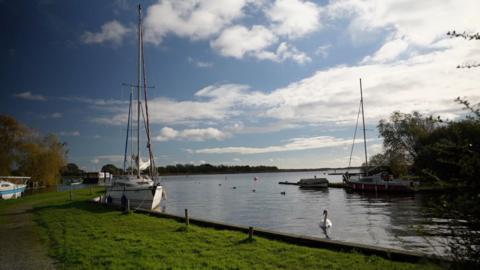 Four small boats are shown moored at the side of a waterway leading into a large broad, with grass banks lined by trees, swans and ducks in the water and a wooded area on the horizon.