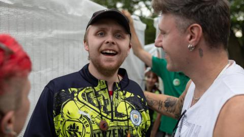 Andre Yarham wears a black and neon yellow top. He is standing with a male friend who has his hand on Yarham's back. Both are smiling.