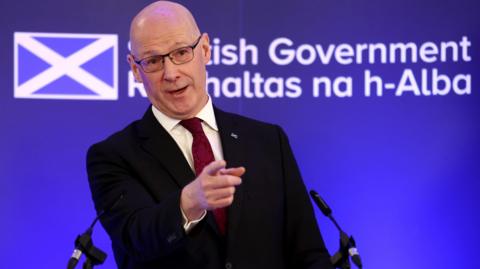 John Swinney points with his right hand while standing at a podium. He is wearing a black suit, cream shirt and a burgundy tie. There is a blue background featuring a Saltire and the Scottish goverment's logo.