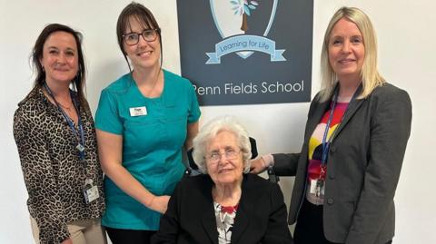 Two women with dark hair and one woman with blonde hair, wearing a grey blazer stand beside a lady with short white hair and glasses who sits in a wheelchair. The signage in the background says 'Penn Fields School'.