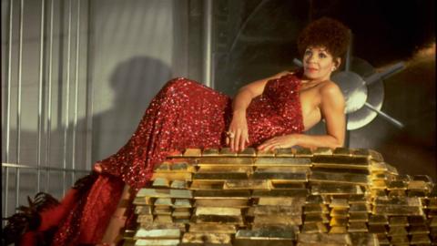 Shirley Bassey lays atop a stack of gold bars