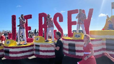 A pink, white and yellow floral float travelling along a road. Large pink letters spell the word Jersey. Three singers are standing on the float holding microphones. The sky is blue.