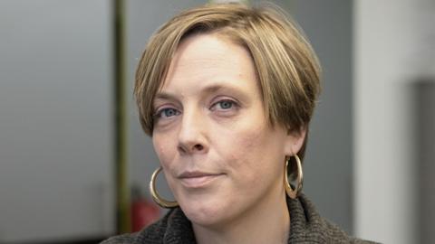 Jess Phillips, with short brown hair and wearing gold hoop earrings. She is facing the camera with a blurred background in an office with grey walls.