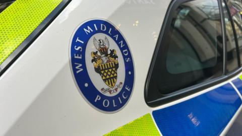 A West Midlands Police emblem stick on the rear part of a white police car in a city centre. 