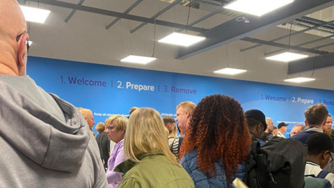 Crowded airport security queue with travellers standing behind green barriers under bright ceiling lights, facing a blue wall sign that reads “Welcome | Prepare | Remove,” while one person in the foreground holds a smartphone.