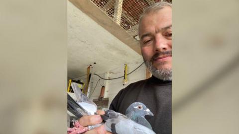 A selfie of Jason Dempsey with one of his pigeons. They are in a shed. Jason has short grey hair and beard and brown eyes. He is wearing a black jumper. 