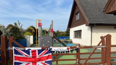There is a soft play area with a climbing frame behind a school gate which has the union flag across it. A sign reads "Welcome to Sark School".