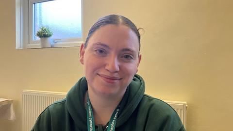 A young woman with brown hair tied back, wearing a green hoodie and sitting at a desk at a Taff Ely Foodbank site. 