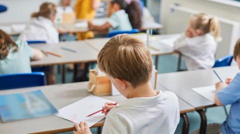 Stock image of children with their back to the camera colour do school work. One main child is visible while the rest are out of focus in the background.