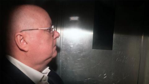 Peter Murrell, who is bald with glasses, photographed in profile in the back of a van. He is wearing a white shirt and black suit jacket, and is illuminated with a flash 
