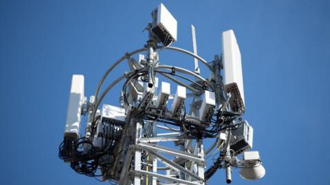 A 5G mobile phone mast, complete with various white boxes on various metal pylons. The sky is blue behind the mast.