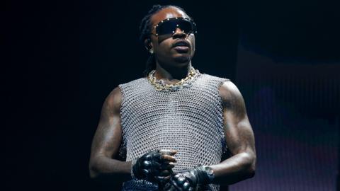 Man in gold and silver chains wears a silver vest top, black gloves and sunglasses while holding a mic