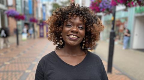 Nyree Hale smiling at the camera wearing a black v-neck t-shirt with shoulder length curly brown hair. She is standing in a town centre which is blurred behind her.