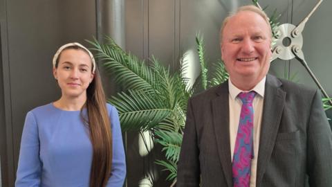 Alex Bulat in a blue dress standing next to Steve Count wearing a grey suit and a pink and blue tie. They are both looking directly at the camera and smiling. There is a green plant behind them. 