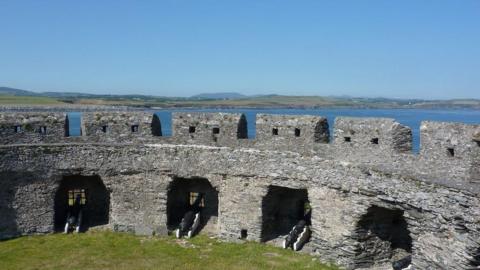 Fort Island from the inside, a stone fort wall arches around, with cannons facing out, there is grass inside, and behind you can see the blue sea and green land of the Isle of Man.