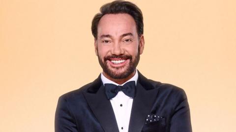 Craig Revel Horwood smiling dressed in a black tuxedo with his arms folded