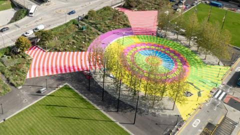 An aerial shot of Station Square with the new artwork. It is shaped in a circle that is edged with tall trees. Concentric circles show vivid patterns of yellow, lime green, fuschia pink and blue. Two red-and-white striped walkways lead out from the circle to two underpasses. Outside the circle are tall flagpoles with various flags, and on either side are large grass squares.