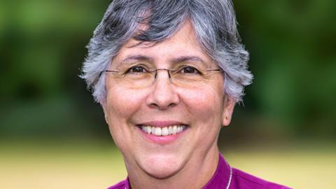 A woman with short hair smiling at the camera. She is wearing a purple top and standing outside.