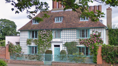 A three-storey village house with a pattern of blue-edged squares on a white background on the front elevation. There is blue door in a white frame. Climbing plants are evident on the facade. There is a small paved area in front of the door with shrubs. There are brick walls either side of the house covered in leaves. A low blue railing separates the property from the road.