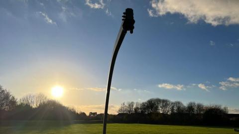 A high black pole for a swing situated in a field. No basket swing hangs from the pole. The field behind is empty and the sun is shining low near trees.