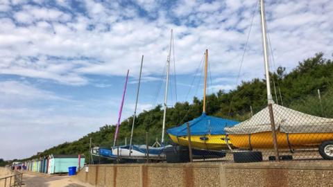 Covered boats are parked on a beach side walk with trees behind and blue sky filled with cloud