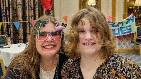 A woman and her teenage daughter smiling at the camera. The woman is wearing pink flamingo sunglasses, a dark coloured cardigan and white top. The teenager has shoulder-length curly hair and is wearing a black and orange pattered shirt. There is colourful bunting and banners in the background.