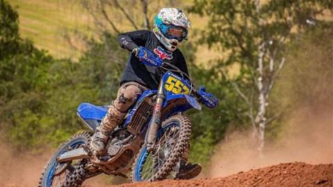 Duncan Powell dressed in black with a blue and white helmet, sat on a blue motorbike covered in mud, on a mud track with trees in the background