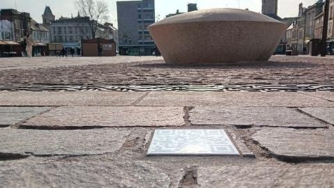 White ceramic tile set amongst porphyry stone paving in the market square. A large circular stone sculpture is visible behind.