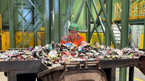 A woman is wearing a green Biffa helmet and an orange high-vis jacket. She is sorting through squashed cans of rubbish.