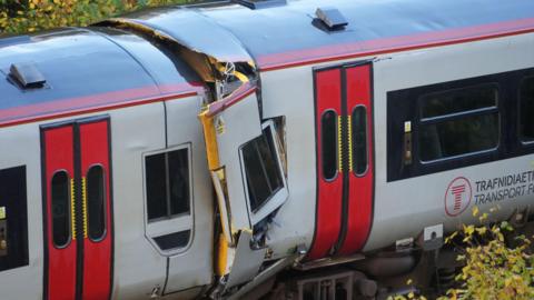 Close-up daytime view of damaged train carriage with window bent sideways and part of outer metal shell hanging off and top of carriage broken open