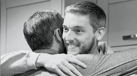Black and white image of two men hugging. One of the men (right) who is facing the camera has a grey beard and hair and is wearing a grey hoodie with the logo Derian Dads on it. He is smiling.