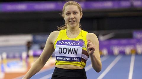 Maddie Down smiles as she runs down a running track. She is wearing a yellow athlete's top with a large name tag on the front. 