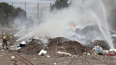 A group of firefighters are extinguishing a fire at an industrial estate using hoses. 