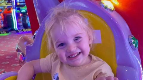 Smiling female toddler with blonde hair in an amusement arcade. She appears to be seated on a ride. She is wearing a t-shirt depicting Minnie Mouse.