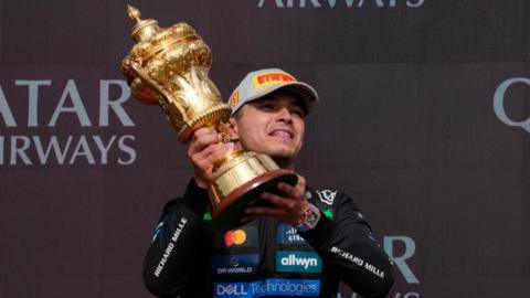 Lando Norris in a black racing suit and cap holding a gold trophy aloft
