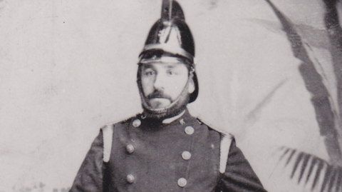 A Victorian photograph of a man with a moustache wearing an old-fashioned firefighters helmet and uniform.