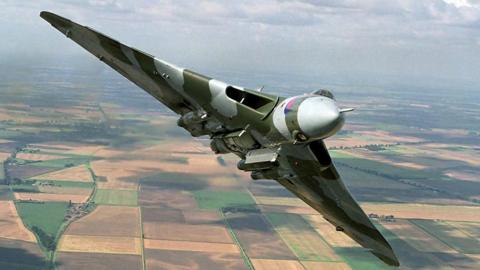 An RAF Vulcan in flight. It has green and grey camouflage paint and is flying over farmers' fields.