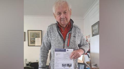 An elderly man with a grey top over a red shirt. He is holding a parking fine and looking into the camera.